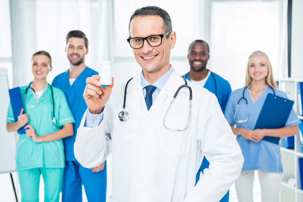 handsome mature doctor holding jar of pills with colleagues standing together on background thumbnail
